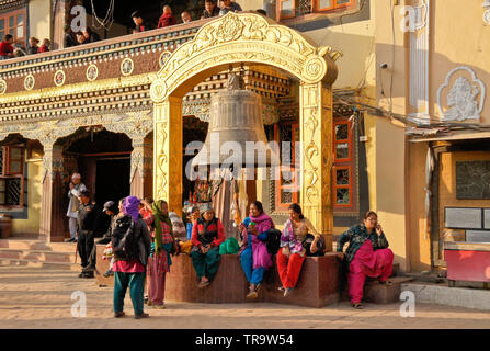 Nepalese people in traditional dress sit beneath big bell in front of Tibetan Buddhist monastery at Boudhanath, Kathmandu Valley, Nepal Stock Photo