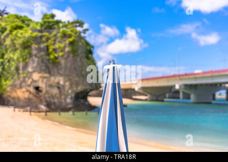 Beach umbrella on the sandy beach Naminoue topped by a huge rock with a Shinto Shrine at the top of a cliff and a highway passing in Naha City in Okin Stock Photo
