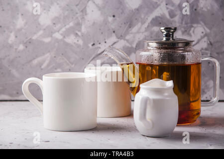 Black tea in a transparent teapot next to white cups and creamer on a gray textural background. Stock Photo
