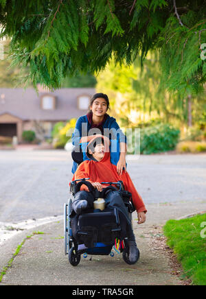 Big sister pushing disabled little brother in wheelchair around neighborhood, laughing and smiling Stock Photo