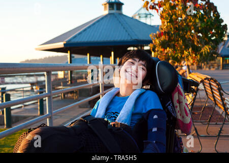 Happy smiling little twelve year old disabled boy in wheelchair outdoors enjoying summer evening at park Stock Photo