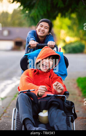 Big sister pushing disabled little brother in wheelchair around neighborhood, laughing and smiling Stock Photo