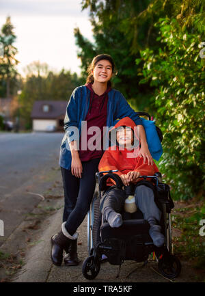 Big sister pushing disabled little brother in wheelchair around neighborhood, laughing and smiling Stock Photo