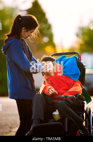 Big sister standing next to disabled little brother in wheelchair while outdoors in neighborhood Stock Photo