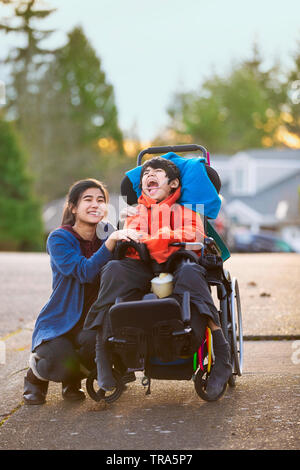 Big sister sitting down next to disabled little brother in wheelchair while outdoors in neighborhood Stock Photo