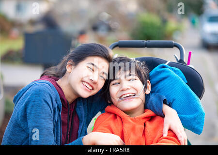 Biracial big sister lovingly hugging disabled little brother in wheelchairoutdoors, smiling Stock Photo