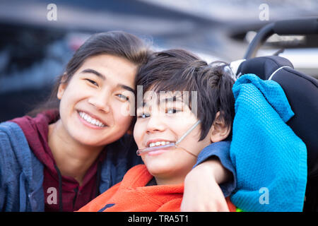 Biracial big sister lovingly hugging disabled little brother in wheelchairoutdoors, smiling Stock Photo