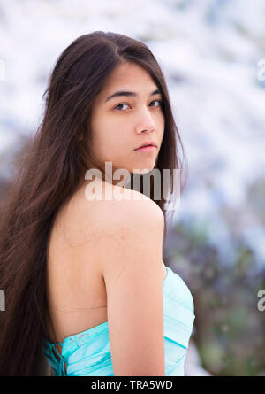 Biracial teen girl or young woman of Asian and Caucasian descent wearing blue dress outdoors in snow looking over shoulder at camera Stock Photo