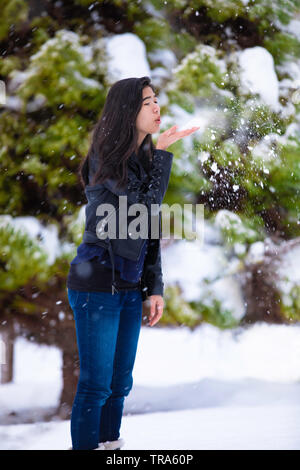 Biracial teen girl standing outdoors in winter blowing snow off hand, with snow covered green pine trees in background Stock Photo