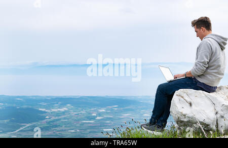 Young man with laptop sitting on top if mountain working online. Cellular network broaband coverage concept. 5G. Stock Photo
