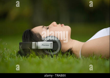 Amazing shot of a modern young Asian woman resting in the grass wearing cool big headphones listening to music