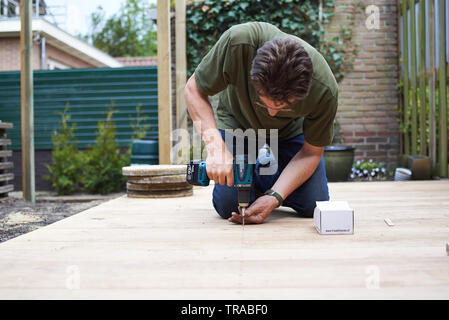 Caucasian carpenter sitting on his knees as is he drilling into wooden plank to make a patio in his back garden Stock Photo