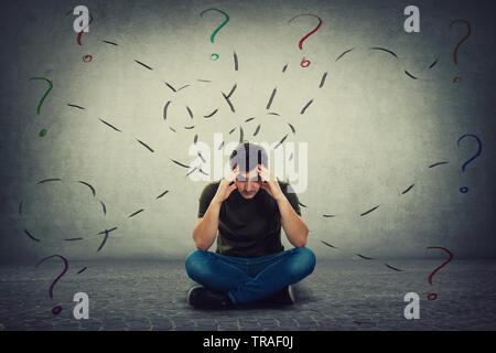 Confused and tired young man seated on the floor keeps hands to forehead looking down thoughtful surrounded by different tasks to solve as having many Stock Photo