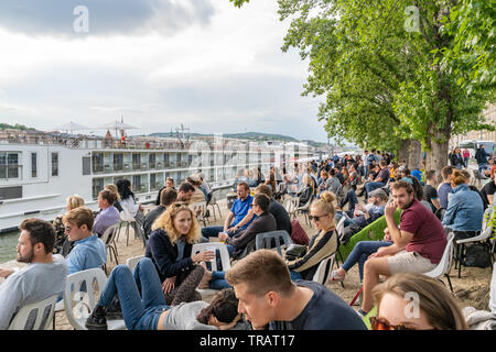 People sitting and socializing with a drink at summer bar on Danube river bank close to a famous Chain Bridge on Pest side of Budapest, Hungary. Stock Photo