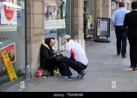 A kind man offers food and drink to a homeless woman begging on a street outside a supermarket with people walking by London England UK  KATHY DEWITT Stock Photo