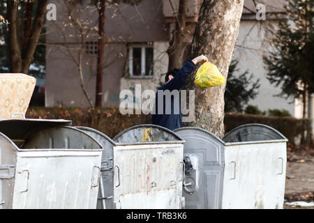 young man throws yellow plastic bag full of garbage in container,  three metal containers on street with building and trees in background Stock Photo