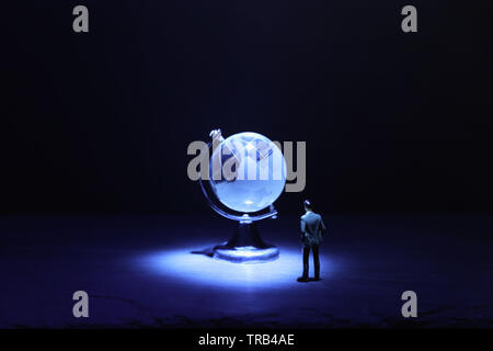 Miniature man looking at Small crystal globe in front of dark and dramatic light.global issues concept Stock Photo
