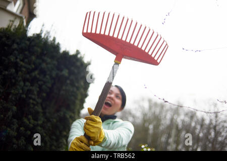 Happy and excited Asian woman wearing yellow rubber gloves and holding a rake during a spring clean session in the garden Stock Photo