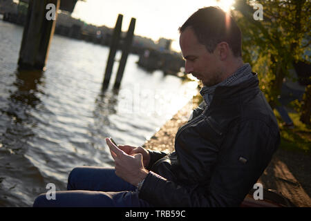 Caucasian man sitting outside in the back garden of a pub along the water using his mobile phone in the city centre of Amsterdam Stock Photo