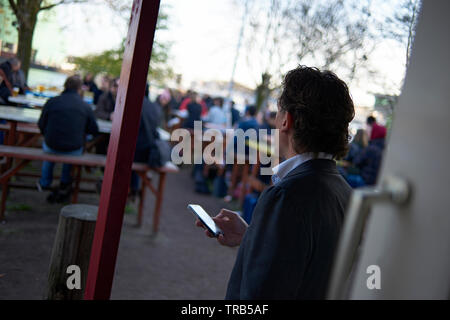 Caucasian man sitting outside in the back garden of a pub along the water using his mobile phone in the city centre of Amsterdam Stock Photo
