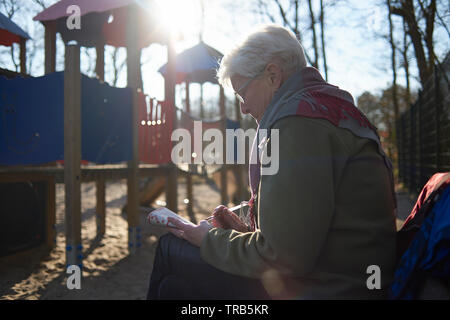 Elderly woman sitting on a bench at a playground reading a book whilst keeping an eye on her grandchild who is playing Stock Photo