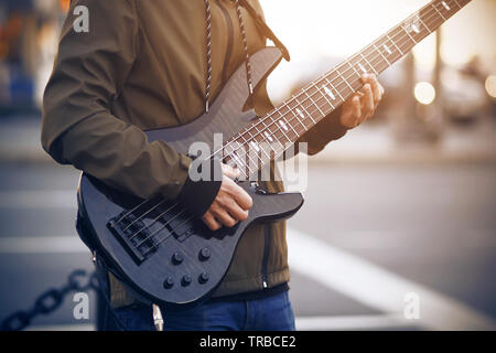 A man dressed in a dark jacket with striped laces plays a beautiful black five-string bass guitar on the street. Stock Photo