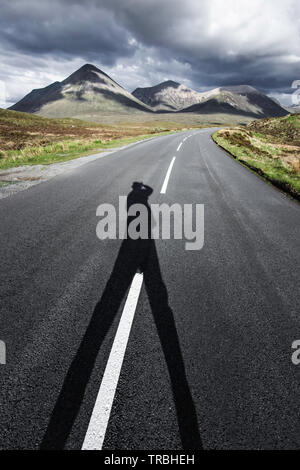 Shadow of person standing on the middle of country road and looking at majestic mountains in background.Travel destination.Diminishing perspective. Stock Photo
