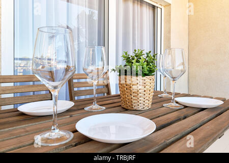 Close up white empty plates wine glasses, wooden table setting at home in outdoors terrace, plant on weave pot no people Stock Photo