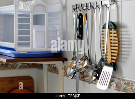 On a tiled wall hanging on hanging rack lot different kitchen stainless utensils in a row Stock Photo