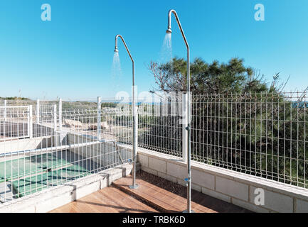 Inside of fenced area two outdoor shower sprinklers is on, water pours on tiled floor outdoors. Photo taken in urbanization near swimming pool Stock Photo