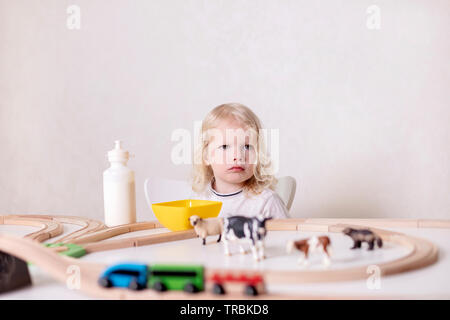 Little cute baby boy (3 years old) drinks milk with cookies and plays in the wooden station train set on the table. Selective focus. Stock Photo