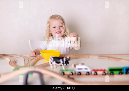 Little cute baby boy (3 years old) drinks milk with cookies and plays in the wooden station train set on the table. Selective focus. Stock Photo