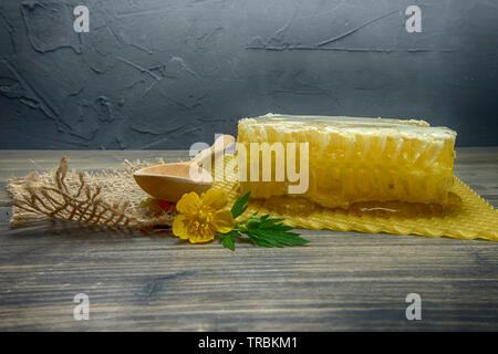 Fresh block of honey comb with honey oozing onto the mat below with a wooden spoon and colorful yellow evening primrose flower, copy space on grey bac Stock Photo