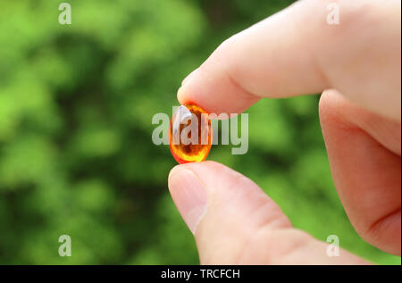 Closeup Man's Fingers Holding a Transparent Supplement Gelcap Against Blurry Green Foliage Stock Photo