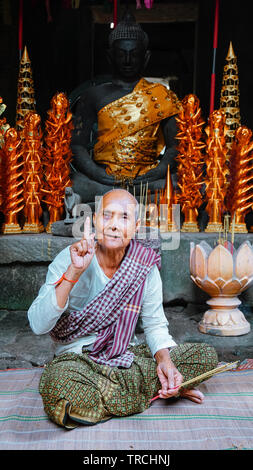 Close-up portrait of a female elderly monk (nun) smiling with one figure pointing up, in front of Buddhist shrine. (Angkor Wat, UNESCO World Heritage Stock Photo