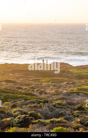 Happy couple enjoying the sunset with an amazing Pacific Ocean view of Big Sur, California coastline from a cliff. Stock Photo