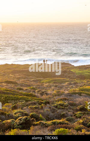 Happy couple enjoying the sunset with an amazing Pacific Ocean view of Big Sur, California coastline from a cliff. Stock Photo