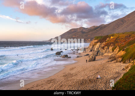 Overlooking a beautiful sandy beach with a woman sitting, reading a book and a man walking along the beach of Big Sur, California. Stock Photo