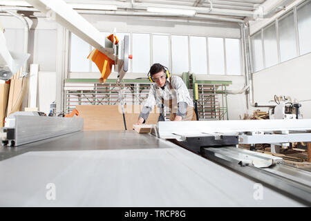 carpenter man work in the joinery, cut a wooden board with circular saw machine, protected with ear muffs and glasses Stock Photo