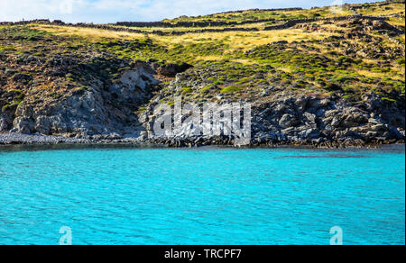 Rocky shore along the clear turquoise blue waters of Dilos showing ancient walls made of stone. Stock Photo