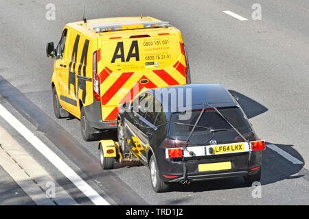 Back view looking down from above on yellow AA Automobile Association business breakdown van towing broken down black car along M25 Motorway Essex UK Stock Photo