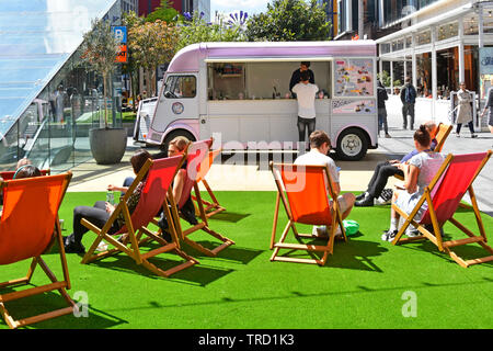 Shoppers mall Westfield shopping centre made over for summer relaxing in deck chairs on artificial grass with street food truck Stratford London UK Stock Photo