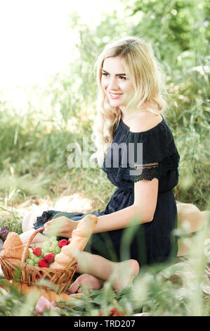 Smiling blond girl having picnic in park. Wearing stylish blue dress. Summer season. 20s. Stock Photo