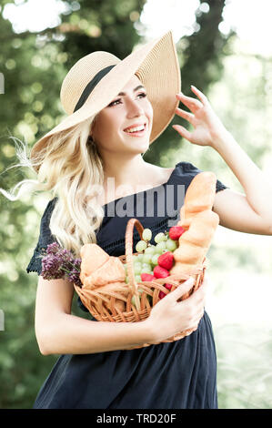 Smiling blonde woman 24-29 year old wearing blue dress and straw hat holding basket with fruits, fresh bread outdoors. Looking forward. 20s. Stock Photo