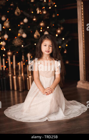 Cute baby girl 4 5 year old wearing stylish dress sitting under Christmas tree with lights in room. Looking at camera. Holiday season Stock Photo Alamy