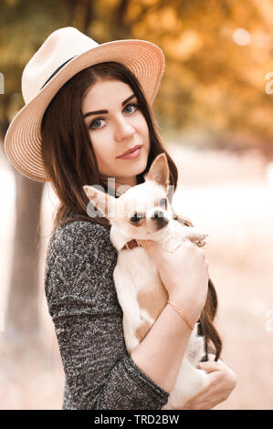Smiling stylish girl 24-29 year old holding chihuahua puppy outdoors. Looking at camera. 20s. Stock Photo