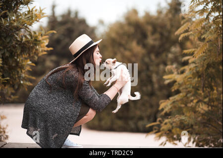 Stylish woman kissing puppy chihuahua outdoors in park. Togetherness. Friendship. 20s. Stock Photo