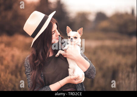 Stylish girl 24-29 year old holding chihuahua puppy wearing summer hat outdoors. Friendship. 20s. Stock Photo