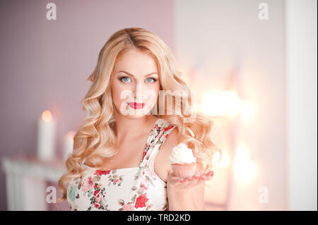 Smiling blonde woman holding cupcake on hand in room over lights. Looking at camera. Stock Photo