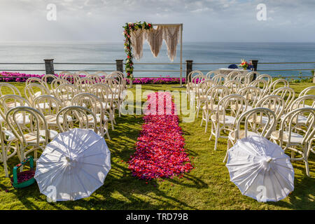 Wedding arch in Boho style near the ocean for ceremony decorated with macrame and fresh flowers. Chairs, umbrellas and pink and red petal aisle. Bali